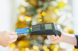 © Alliance - A close-up captures a hand inserting a blue credit card into a reader, held by another hand. The blurred, warm lights in the background suggest a festive retail transaction during the holiday season.