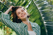 © SHOTPRIME STUDIO - Young woman smiling outdoors, wearing a striped green shirt, captured amid bright sunlight and lush tropical foliage for a fresh, natural portrait.