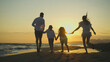 © Videophilia - Family of four—parents and two children—running hand in hand along the sandy shoreline at golden hour, silhouetted against a warm orange sunset over the sea