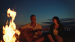 © Videophilia - Young man plays acoustic guitar beside a warm bonfire as his girlfriend smiles, sharing a cozy, romantic beach moment at dusk during a relaxed summer evening getaway