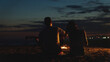 © Videophilia - Couple sitting on a sandy beach at night, a man playing an acoustic guitar by a warm bonfire, with a woman sitting close, enjoying music and the twilight ocean view