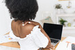 © etonastenka - Close-up of African American woman sitting at desk with laptop, feeling back and neck pain from long hours and poor posture. Concept of home office, remote work, and health problem.