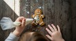 © Таня Андреева - Top-down view of a child's hands using a piping bag to decorate a gingerbread man with white icing. Colorful sprinkles are on a rustic wooden table.