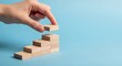 © Vamika - Hand placing wooden block on staircase of blocks, symbolizing progress, growth, development, achievement, and step-by-step success in business, career, and personal goal attainment on blue background.