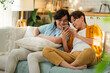 © Yingyaipumi - Asian male couple smiling and relaxing together on a cozy sofa, browsing social media or shopping online on a smartphone.Concept of LGBTQ+ relationship, domestic life and digital connection.