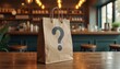 © Pete - A brown paper bag with a question mark stands on wooden table. The cafe background is blurred. The photo concept is mystery expectation delivery takeaway.