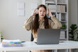 © Wasan - Smiling young Asian businesswoman wearing headphones and working from home using laptop, sitting at desk with documents and pen, in home office.