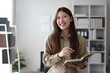 © Wasan - Cheerful Asian businesswoman holding a notebook and smiling in the office.