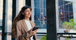 © serg - Confident and smiling young businesswoman walking outside modern office building, smiling while using smartphone, checking messages, browsing apps, staying connected on the go, outdoor