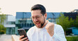 © serg - Attractive young man wearing white shirt holding phone and celebrating success, looking at cellphone screen, raising fist with excitement at modern glass building background outdoors