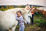 Equine assisted therapy with child and horses on animal farm. Little girl embracing white pony horse while her mother is watching them