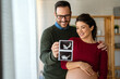 © NDABCREATIVITY - Happy young couple, pregnant woman, holding and showing ultrasound scan picture at home