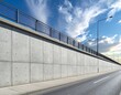 © Baladi - Modern highway construction with a long concrete retaining wall, metal railing, asphalt road, and a cloudy blue sky