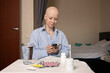 © yta - Bald woman after chemotherapy using smartphone at table with pills and water, concept of buying cancer drugs online and digital healthcare support.