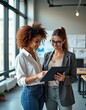 © Viktor - Two businesswomen review tablet content in office. Females discuss project plan. Colleagues collaborate on work using tech. Professionals brainstorm. Modern workspace with diverse team sharing ideas.