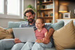 © Stockphotodirectors - Father and daughter enjoy quality time together, smiling and sharing laughter as they explore content on a laptop while sitting on a comfortable sofa in a warm living room.
