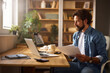 © Stockphotodirectors - A man is engaged in work at a modern home office, seated at a wooden desk with a laptop and papers in front of him while surrounded by houseplants and shelves.