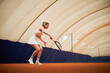 © DragonImages - Caucasian young adult woman playing tennis on indoor clay court, holding racket and preparing to hit ball, focused on action, athletic movement under large dome structure