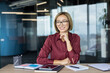 © Liubomir - Young businesswoman in glasses smiling thoughtfully with hand on chin at her desk in a modern office, planning and focused on strategy with tablet and notebook nearby