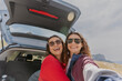 © olezzo - Two girlfriends taking selfie through smart phone while sitting in car trunk with mountains view