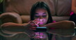 © Marco - Young girl resting her head on arms while using smartphone on glass table, focused and engaged with screen in dim indoor setting, glowing light on face
