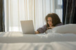© shine - Relaxed asian middle aged woman sitting on comfortable bed at home or hotel and using laptop looking at screen working in internet, enjoying a new day, feeling rested and refreshed. Copy space