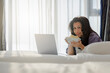 © shine - Lifestyle image of A middle-aged woman using social media on laptop laying on bed in morning, holding pop corn plate, looking to camera and Smile. Wireless connection in hotel or home.