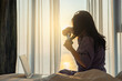 © shine - Beautiful model woman long hair using wireless curling iron or hair straightener making curls in the morning on bed with laptop with large window Bright sunlight. Healthy lifestyle, Morning routine.