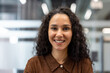 © Liubomir - Young curly-haired businesswoman smiling confidently at camera in a modern office headshot, conveying positivity, professionalism and successful leadership in a casual corporate setting