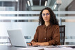 © Liubomir - Young confident businesswoman wearing glasses and a brown shirt, looking at the camera while typing on a laptop, sitting at a modern office desk with documents