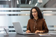 © Liubomir - Professional woman focused on a laptop during an online video conference in a modern office, wearing wired earphones while participating in a virtual meeting and working remotely