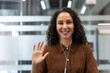 © Liubomir - Smiling young woman with curly hair wearing wired earphones, waving hand and looking at camera during an online video call from a modern office environment