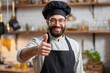 © Vitalii Shkurko - A chef with glasses and a beard smiles and gives a thumbs up in a warm kitchen filled with cooking tools. He wears an apron and a chef hat, showing confidence and satisfaction