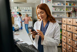Smiling female pharmacist using cell phone while working in pharmacy.