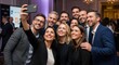 © NanieStudio - Happy business team taking a group selfie with a phone at a corporate event. Colleagues celebrating success together at a gala. Team building, fun, networking at a conference.