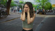 © Krakenimages.com - Hispanic young woman cups ears with hands amid bustling city street traffic and passing bicycles; noise frustration overwhelm.