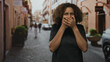 © Krakenimages.com - Young african american woman covering mouth with both hands on a cobblestone city street, wide eyes and casual black shirt; surprise discovery.
