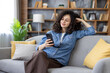 © Liubomir - Young woman with curly hair sitting on a gray sofa at home, smiling and relaxing while scrolling social media on her smartphone, enjoying cozy digital leisure and connection