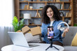 © Liubomir - Young woman smiling, presenting new wireless headphones from an open cardboard box to a smartphone on a tripod, creating content for her social media vlog