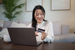 © matinee - Young woman making online payment or e-commerce purchase with a credit card in a cozy living room.