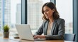 © Fabio - Successful smiling businesswoman working on a laptop in a modern office. Confident female professional or entrepreneur typing on a computer at her desk with a city view background.