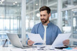 © Liubomir - Business man with a beard smiling while reviewing financial documents, managing paperwork, and using a laptop at his desk in a modern corporate office, symbolizing planning and success