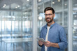 © Liubomir - Young businessman wearing glasses and a blue shirt holding a digital tablet and smiling at the camera in a modern glass office interior, symbolizing business, technology, and success