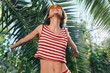 © SHOTPRIME STUDIO - A cheerful woman enjoys sunny outdoors in a tropical setting. She wears a red and white striped top, smiles brightly, and opens her arms, radiating carefree energy and summer vitality.