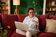 © Stockphotodirectors - A woman sitting on a red sofa is happily using her laptop to shop online. She holds a credit card in her hand and enjoys a relaxed atmosphere in her living room.