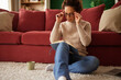 © Stockphotodirectors - A woman in a cozy sweater sits on a rug with her notebook, adjusting her glasses as she contemplates her notes. The living room is warm and inviting, decorated with cushions and plants.