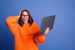 © deagreez - Excited young woman holding laptop in orange sweater against purple backdrop, expressing surprise and joy in casual style.