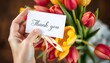 © Husen - A hand holding a thank you card in front of a bouquet of colorful tulips.