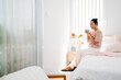 © artitwpd - Woman enjoying a hot drink on a bright minimalist bedroom, sitting on bed near window and flowers, peaceful morning home scene.