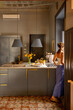 © rh2010 - Woman enjoying a peaceful morning in a stylish Sicilian kitchen with vintage tiles, gray cabinets, and yellow flowers. Cozy Mediterranean home atmosphere